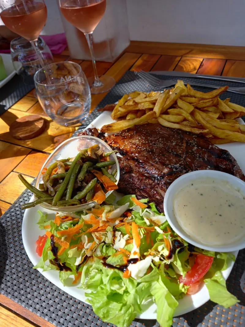 Une entrecôte grillée avec ses frites, sa salade, ses haricots verts et sa sauce, sur une table en teck
