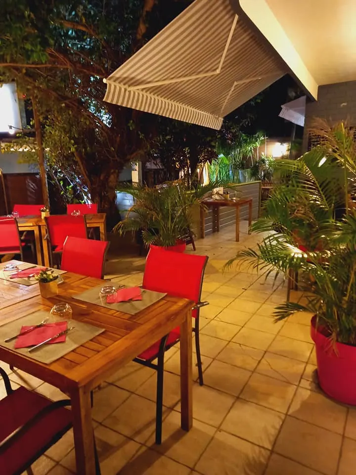 Les tables en teck et les chaises framboise de la terrasse, le soir, entre les palmiers en pot
